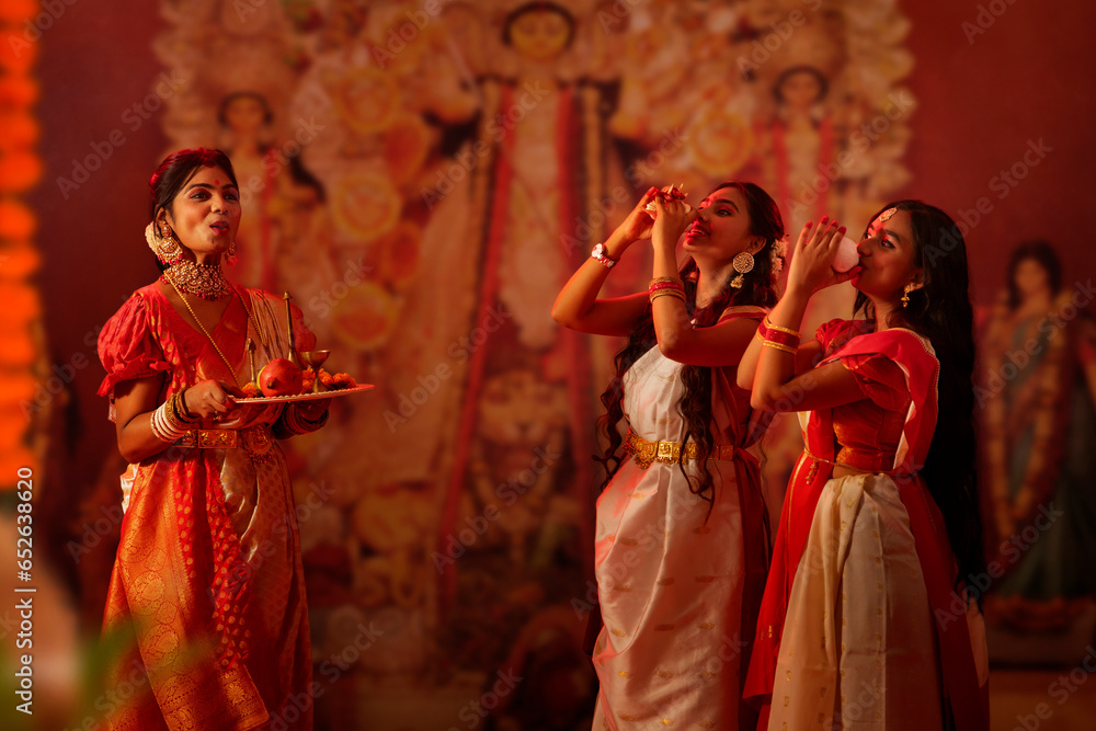 Two Bengali sister playing conch on the occasion of Durga Puja while mother holding a puja thali