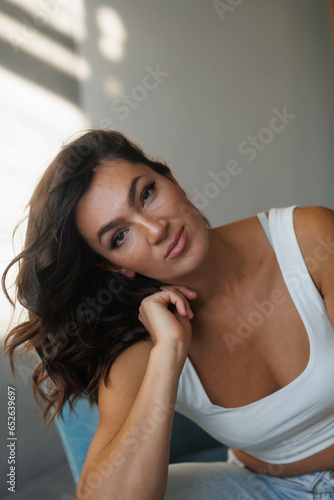 Beautiful brunette woman in white top and blue jeans sitting. Girl smile, happy. Portrait of young pretty woman. Smile and freshness. White background. Light from window, shadows