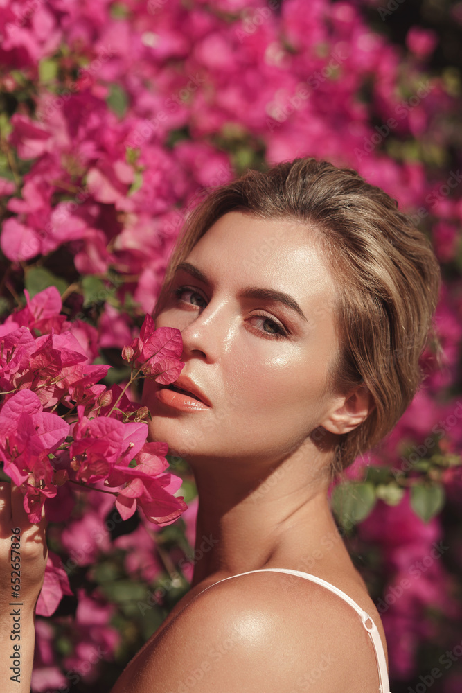 Naklejka premium Beautiful blonde woman with smooth skin and natural makeup, as she poses against a backdrop of a bush with delicate pink flowers