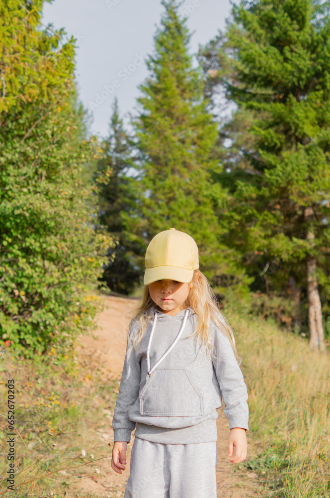 A cute little girl in a yellow cap with blonde natural hair and casual clothes walking along a path in a beautiful forest in the autumn, vertical photography