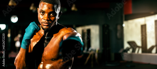 Boxing fighter shirtless posing, african boxer punch his fist in front of camera in aggressive stance and ready to fight at gym with kicking bag and boxing equipment in background. Spur