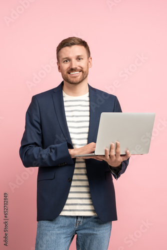 Positive man trader, crypto investor, entrepreneur, financier holds laptop on pink background. Bearded attractive smiling guy wearing jacket, jeans looking at camera on advertisement banner poster.