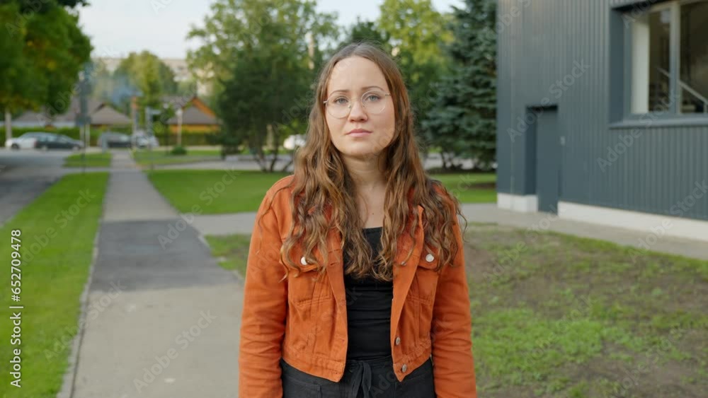 Woman in Orange Jacket Stands on Sidewalk, Gazing Away with a Somber Expression Outside an Office Woman in Orange Jacket Stands on Sidewalk, Gazing Away with a Somber Expression Outside an Office
