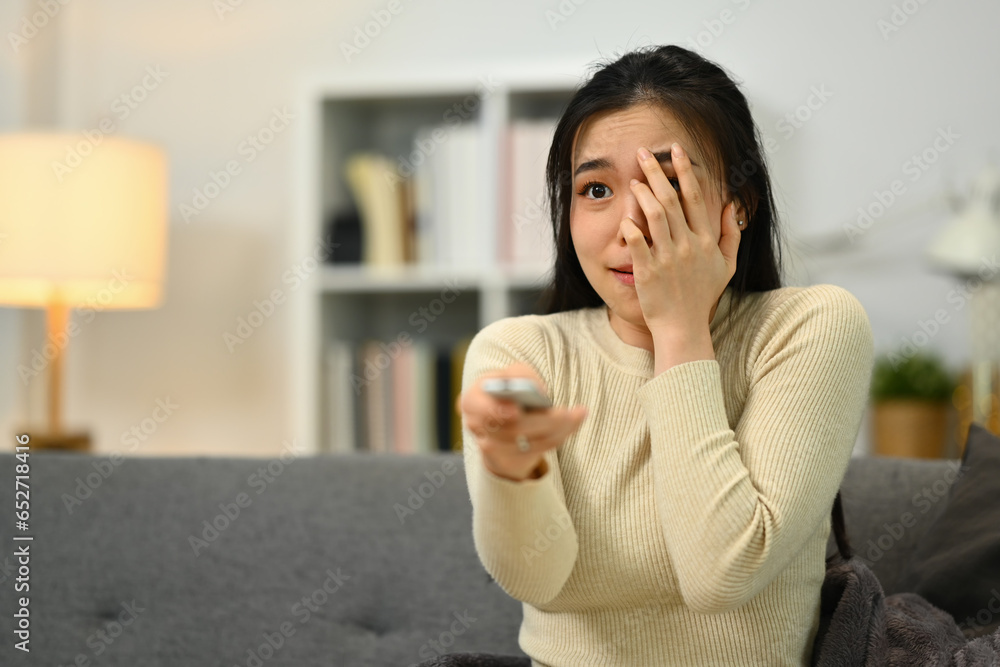 Scared young asian woman with remote control watching scary movie at home. Entertainment and hobby concept Scared young asian woman with remote control watching scary movie at home. Entertainment and hobby concept