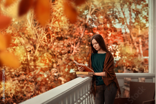 Wallpaper Mural A girl reads a book on the balcony of her house. She is thoughtful, dressed in a green sweater and brown shawl. Season of the year is autumn. There are many autumn trees near the house. Torontodigital.ca