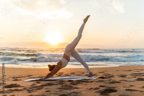 Young woman in sportswear doing yoga exercise with raised leg outdoors, having workout on sea beach at sunset, side view