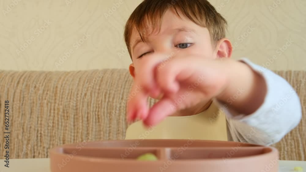 The baby is happily eating fruits and berries, sitting at the table with a bib on. Kid boy aged two years (two-year-old) The baby is happily eating fruits and berries, sitting at the table with a bib on. Kid boy aged two years (two-year-old)