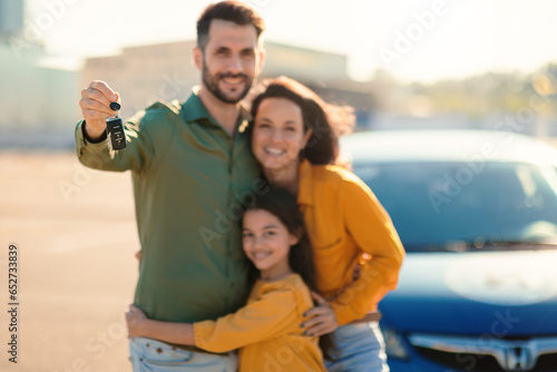 Excited family of three showing new car key, standing near luxury auto outdoors and smiling at camera, selective focus