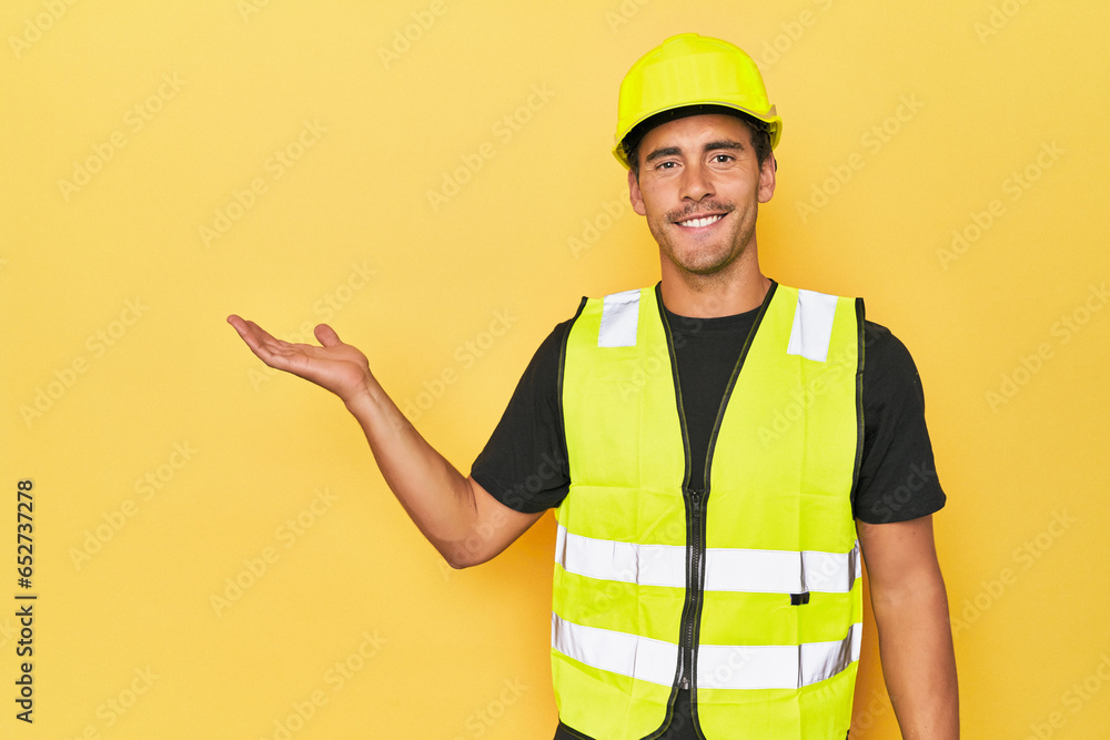 Latino worker in yellow vest and helmet showing a copy space on a palm and holding another hand on waist.