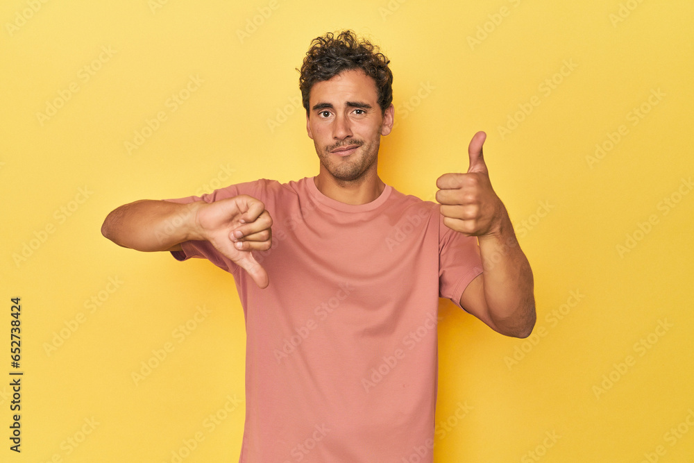 Young Latino man posing on yellow background showing thumbs up and thumbs down, difficult choose concept Young Latino man posing on yellow background showing thumbs up and thumbs down, difficult choose concept