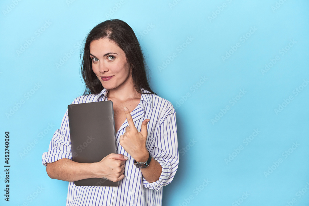 Young businesswoman holding laptop on blue pointing with finger at you as if inviting come closer. Young businesswoman holding laptop on blue pointing with finger at you as if inviting come closer.