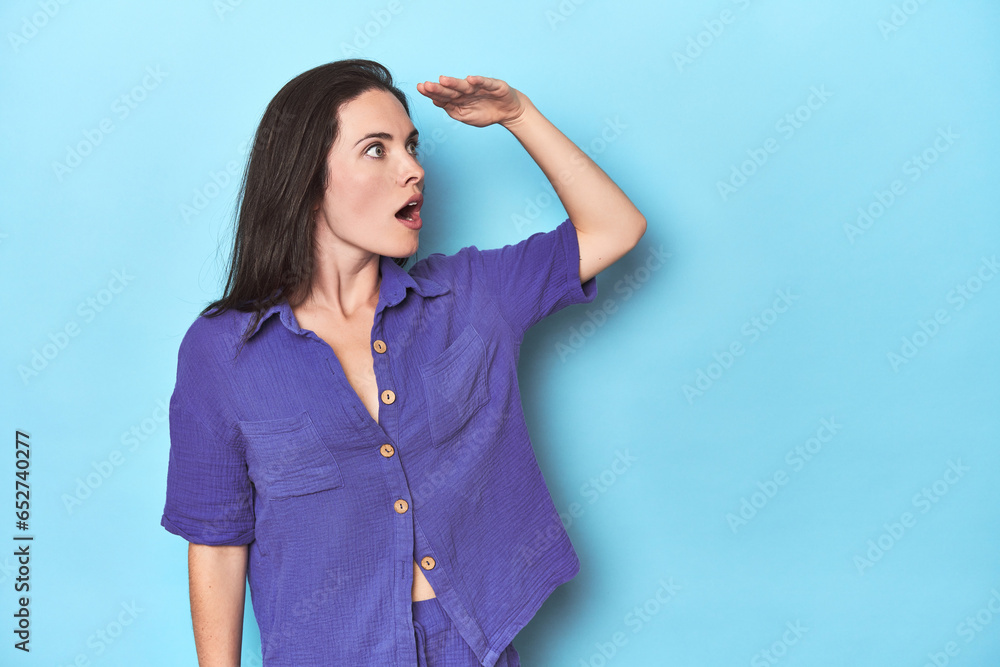 Young caucasian woman on blue backdrop looking far away keeping hand on forehead. Young caucasian woman on blue backdrop looking far away keeping hand on forehead.