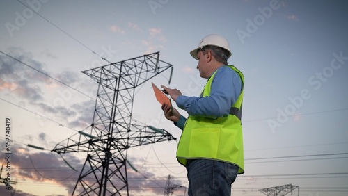 Engineer writes report on tablet looking at power transmission lines in field © SUPER FOX