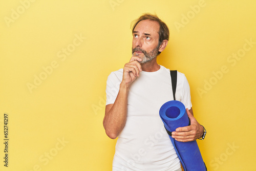Man with yoga mat in yellow studio looking sideways with doubtful and skeptical expression.