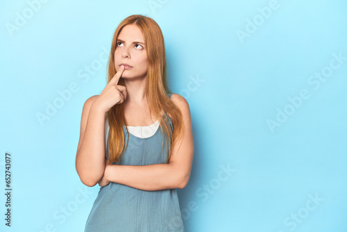 Redhead young woman on blue background looking sideways with doubtful and skeptical expression.