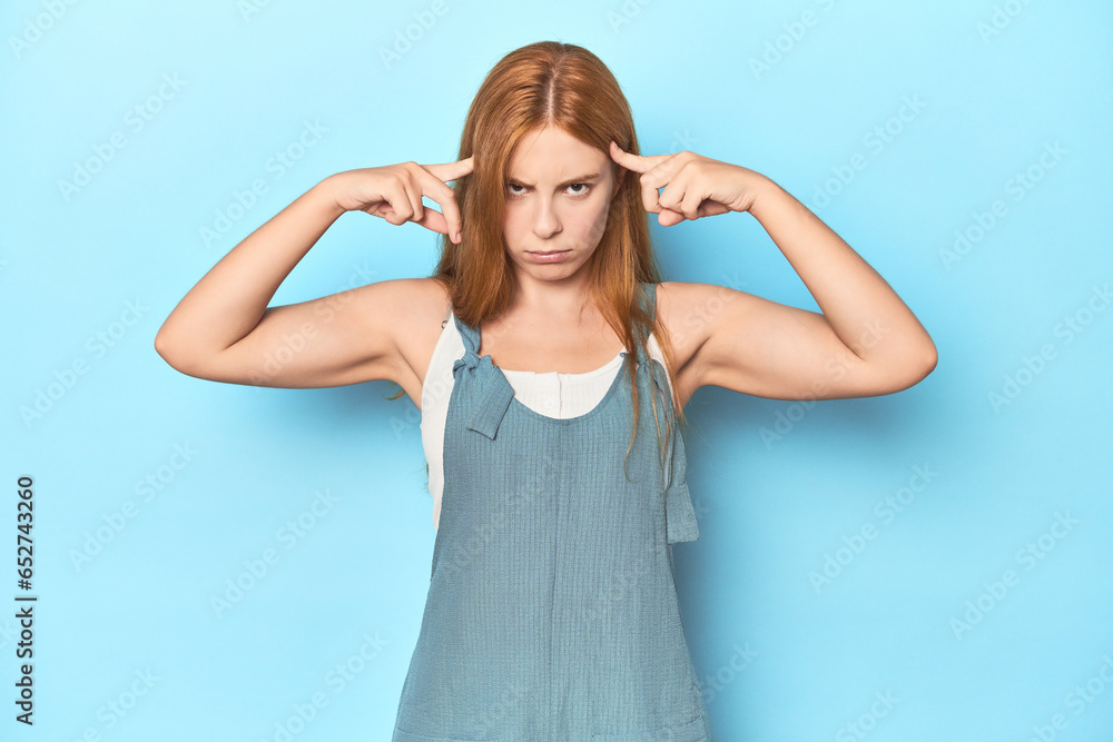 Redhead young woman on blue background focused on a task, keeping forefingers pointing head. Redhead young woman on blue background focused on a task, keeping forefingers pointing head.