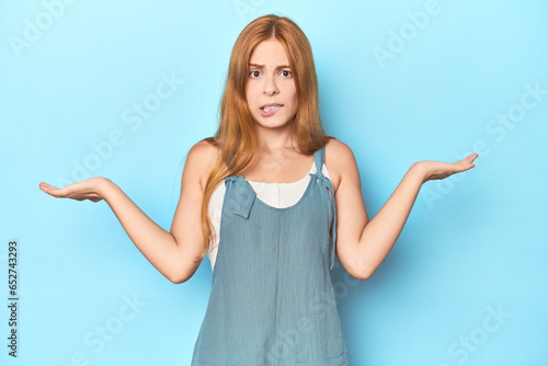 Redhead young woman on blue background confused and doubtful shrugging shoulders to hold a copy space. © Asier