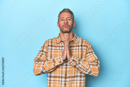 Middle-aged caucasian man on blue backdrop praying, showing devotion, religious person looking for divine inspiration.