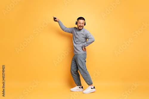 Happy Asian Man Wearing Wireless headphones Dancing Over Yellow Studio Background photo