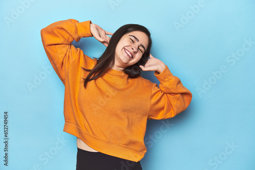 Young Caucasian woman on blue backdrop stretching arms, relaxed position. © Asier