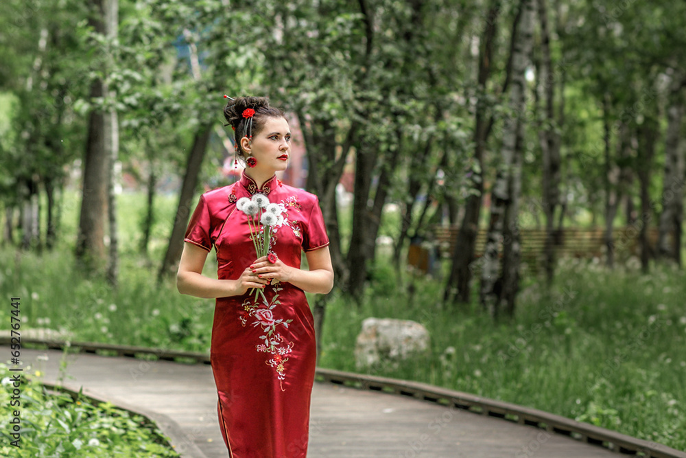 A young Asian woman walks in national clothes in the park. Chinese woman in a red traditional dress holds a bouquet of white dandelions in her hands, close-up portrait. Blows away the fluffs