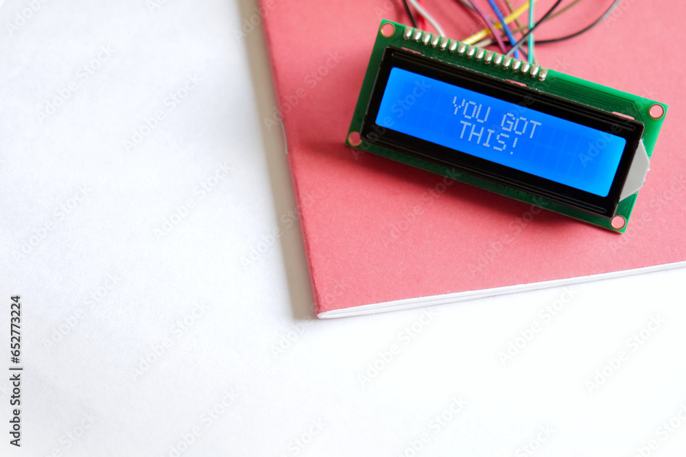 A glowing blue LCD Display shows the positive affirmation 'You Got This'. The display is placed on a red school exercise book, on a white paper background. A glowing blue LCD Display shows the positive affirmation 'You Got This'. The display is placed on a red school exercise book, on a white paper background.