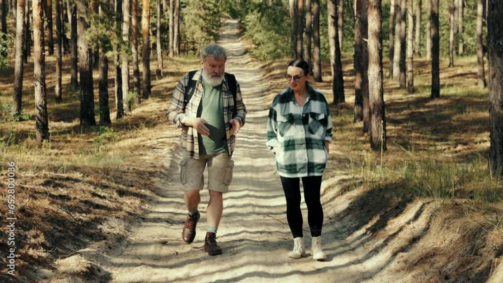Senior people, man and woman walking in forest on warm sunny day, talking, spending good active time together. Concept of leisure time, active lifestyle, hobby, weekends, nature, health Senior people, man and woman walking in forest on warm sunny day, talking, spending good active time together. Concept of leisure time, active lifestyle, hobby, weekends, nature, health