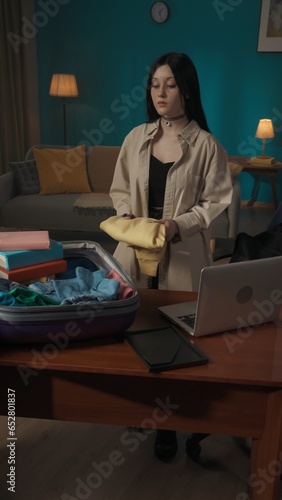 Vertical photo, shot of a teenager, teen, young woman packing her belongings in a suitcase on a table. She is moving out or going on a vacation, holiday.