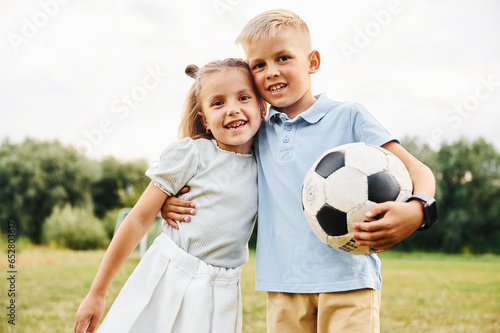 Holding soccer ball and embracing each other. Boy and girl are together having fun on the field at daytime