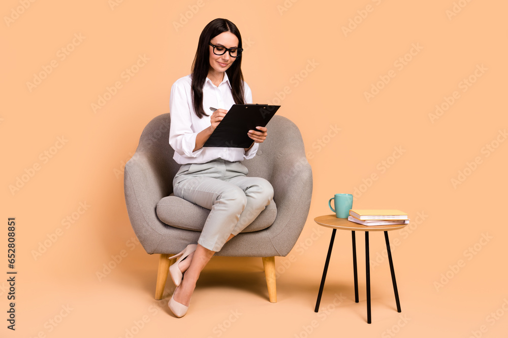 Full body portrait of classy gorgeous financier lady sitting comfort soft chair hold pen write clipboard isolated on beige color background Full body portrait of classy gorgeous financier lady sitting comfort soft chair hold pen write clipboard isolated on beige color background