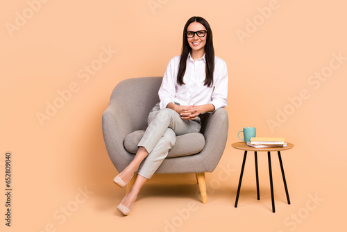 Full size portrait of pretty positive classy girl sitting comfy soft chair coffee table isolated on beige color background