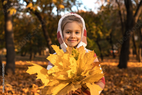 Portrait of little happy girl holds bouquet of yellow maple leaves in autumn park in evening at sunset. Leisure  weekend outdoors