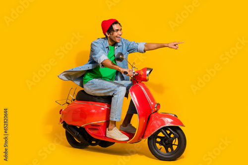 Full body photo of attractive young man driving scooter point empty space wear trendy jeans clothes isolated on yellow color background © deagreez
