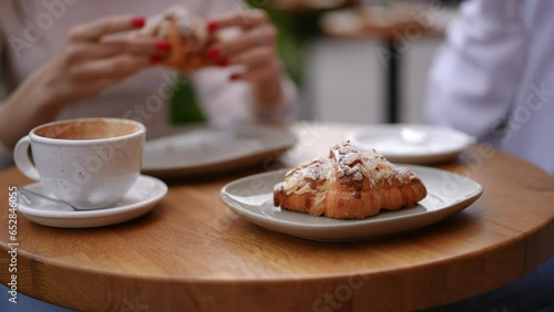 Close-up coffee cup and croissant with blurred female hands at background. Unrecognizable Caucasian young women eating dessert drinking hot drink in cafe on urban city street outdoors