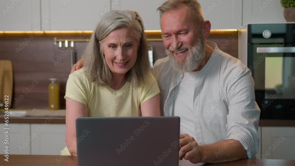 Middle aged family talk use laptop at kitchen browsing social networks book tickets shopping online food delivery smiling Caucasian man and woman buy fresh products old retired couple with computer