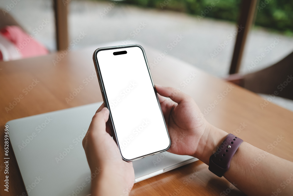 A woman is sitting in a cafe, holding a blank screen mock-up mobile phone in one hand and a coffee cup in the other. A woman is sitting in a cafe, holding a blank screen mock-up mobile phone in one hand and a coffee cup in the other.