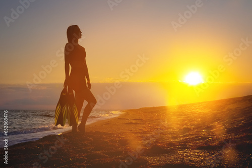 girl diver holds fins in her hands. Summer vacation. Female sexy body silhouette during sunset on vacation after swimming with fins