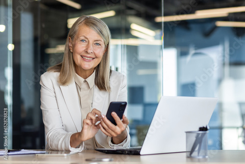 Portrait of mature gray-haired businesswoman inside office at workplace, woman boss smiling and looking at camera, holding phone in hands using smartphone app, sitting at table with laptop. photo