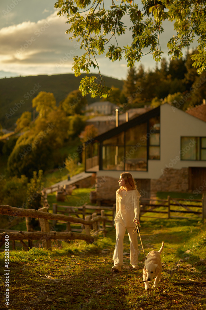 Fototapeta premium Chica pasea con cachorro golden retriever en jardín de casa montañesa. Momentos felices en un paisaje majestuoso.