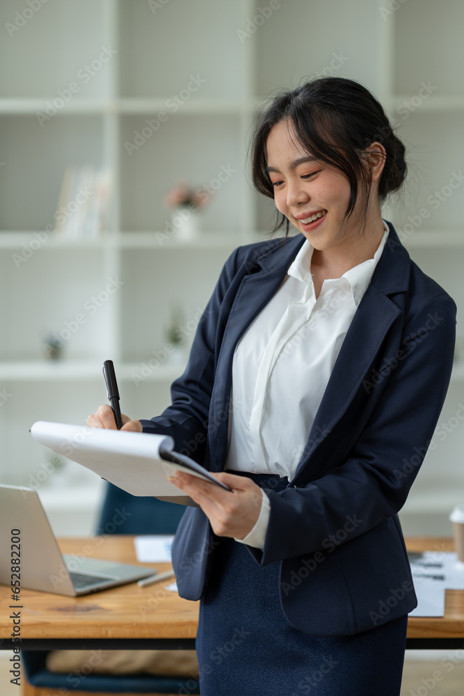 Asian businesswoman standing working with laptop computer, mobile phone, tablet to contact, chat, search internet, news, email, chat while working on finance, marketing, accounting data in office. Asian businesswoman standing working with laptop computer, mobile phone, tablet to contact, chat, search internet, news, email, chat while working on finance, marketing, accounting data in office.