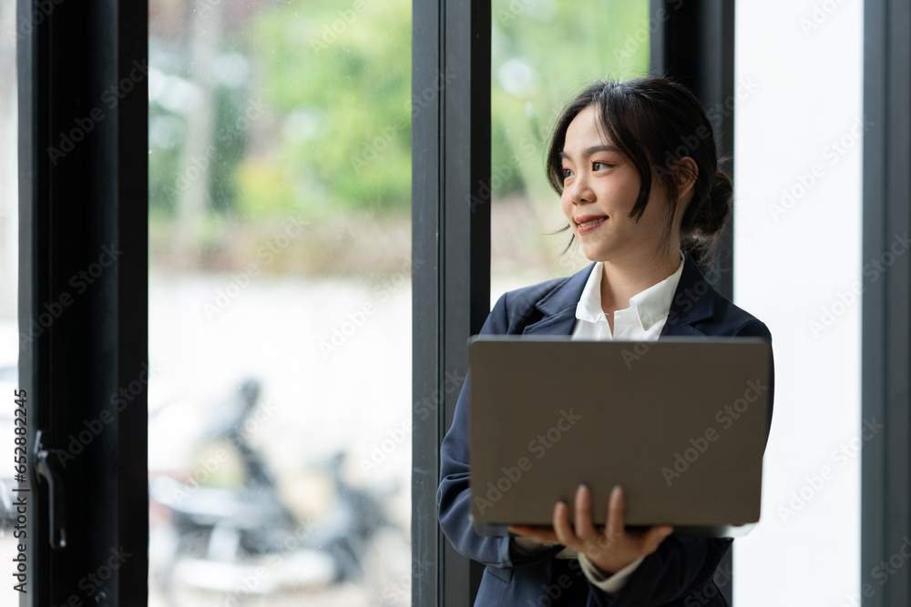 Asian businesswoman standing working with laptop computer, mobile phone, tablet to contact, chat, search internet, news, email, chat while working on finance, marketing, accounting data in office. Asian businesswoman standing working with laptop computer, mobile phone, tablet to contact, chat, search internet, news, email, chat while working on finance, marketing, accounting data in office.