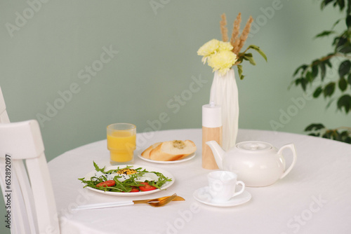 Healthy breakfast of fried egg with tomatoes and green on white plate, orange juice, bread, teapot and cup of hot tea on white table.