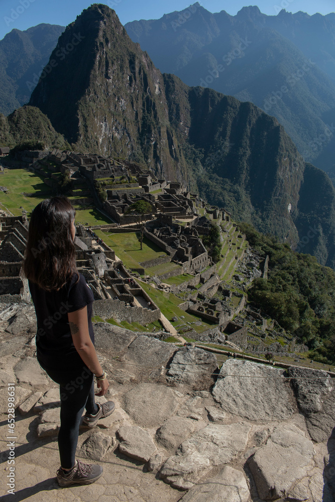Obraz premium Young woman with long flowing hair, wearing sportswear, with her back turned looking at the archeological site of Machupicchu, Peru. 