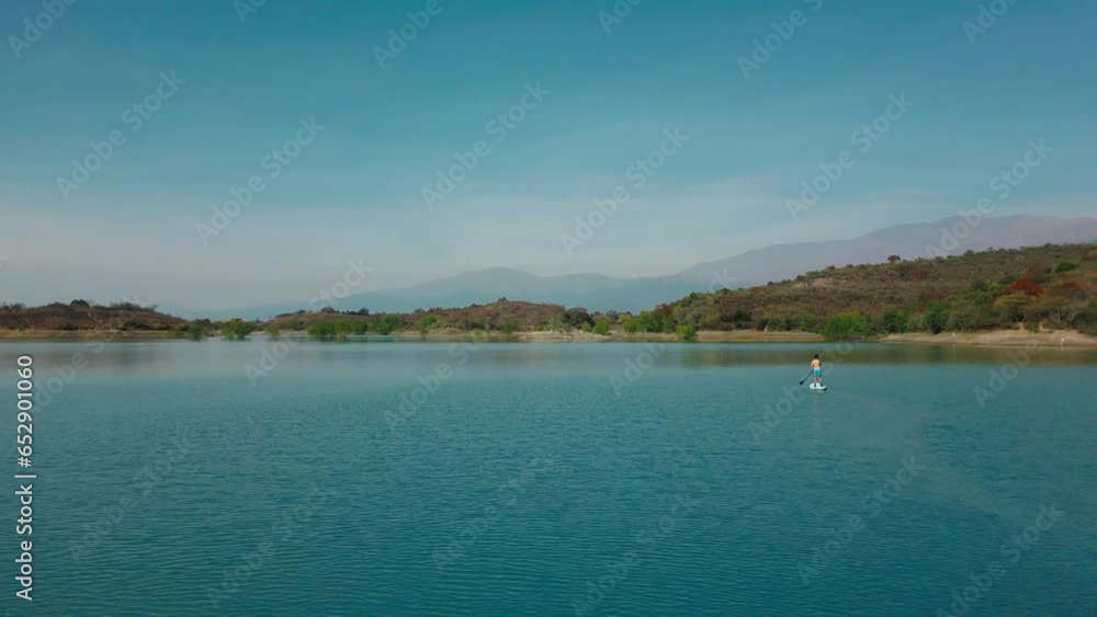 Aerial drone flying over a man doing stand up paddle in a blue water dike lake with mountains landscape Aerial drone flying over a man doing stand up paddle in a blue water dike lake with mountains landscape