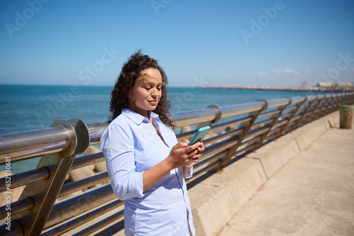 Beautiful ethnic woman in blue casual shirt checking mobile app on her smartphone, smiling, enjoying beautiful sunny day