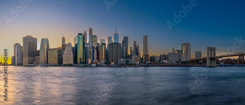 Panoramic cityscape about lower Manhattan s skyscrapers.  The photo taken from brooklyn bridge park. Amazing blue hour view and the sundown on the left side  .Old pier s columns on the foreground.