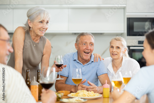 Φωτογραφία Happy carefree older man and women gathering around kitchen table, sipping alcoholic drinks with snacks, cheerfully chatting, laughing and sharing stories