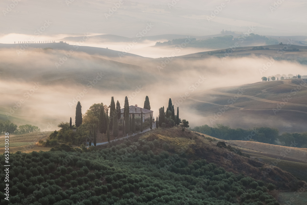 Atmospheric sunrise mist through the valley of Val d'Orcia and a rustic farmhouse in the scenic countryside landscape and rolling hills of rural Tuscany, Italy.