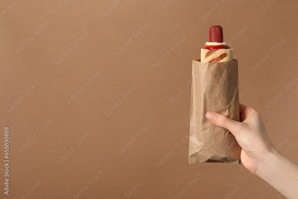 Woman holding paper bag with delicious french hot dog on brown background, closeup. Space for text - obrazy, fototapety, plakaty 