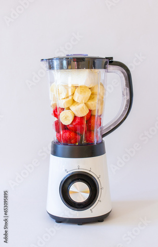 White mixer with fruits and berries, banana, strawberry on gray background, making smoothie, vertical photo
